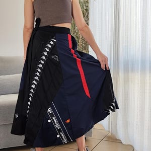 Peut inclure: Une jupe portefeuille noire et bleue avec une bande rouge et des logos Kappa blancs. La jupe est faite d'un tissu léger et a une silhouette longue et fluide.
