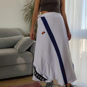 May include: White wrap skirt with blue and red stripes and a Kappa logo. The skirt is made of a sporty material and has a long, flowing silhouette.