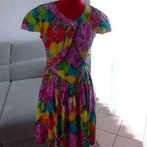 Può includere: Un abito a stampa floreale colorato con una giacca abbinata a maniche corte. L'abito ha un corpetto aderente e una gonna ampia. Il tessuto è una miscela di cotone leggero.