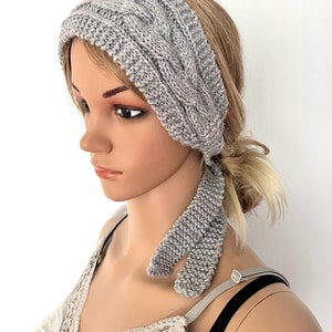 Puede incluir: Una diadema de punto gris con un patrón de punto trenzado y una larga corbata. La diadema se lleva en un maniquí con el pelo rubio.
