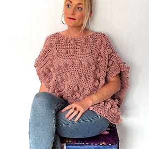 May include: A woman wearing a pink crocheted top with a textured bobble stitch pattern, blue jeans, and coral-colored heels. She is sitting on a colorful striped stool with wooden legs.