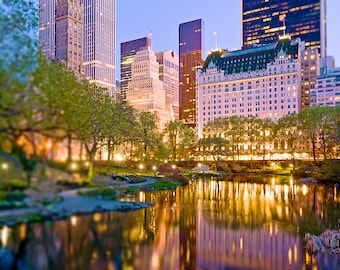 Puede incluir: Una vista de un horizonte urbano al atardecer con un estanque reflectante en primer plano. Los edificios están iluminados y se reflejan en el agua. Hay árboles y arbustos en primer plano.