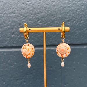 May include: A pair of gold hoop earrings with seashell-shaped pendants. The pendants are cream-colored with orange patterns and feature a small pearl drop. The earrings are displayed on a gold stand against a blue background.