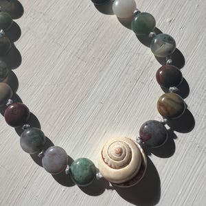 May include: A beaded necklace featuring a large, cream-colored seashell pendant. The necklace is strung with a variety of colorful, round beads in shades of green, brown, and gray. The beads are connected by a white string.