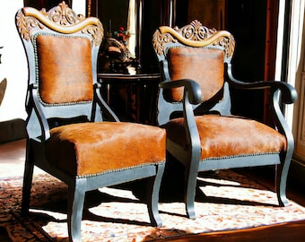 May include: Two brown leather armchairs with ornate wooden frames and black painted legs. The chairs are upholstered in brown cowhide with a distressed finish.