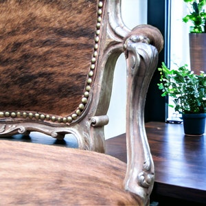 Pu&ograve; includere: Una poltrona in stile vintage con rivestimento in pelle di mucca marrone e bianca. La sedia ha una struttura in legno intagliato con finitura argentata e un bracciolo decorativo.