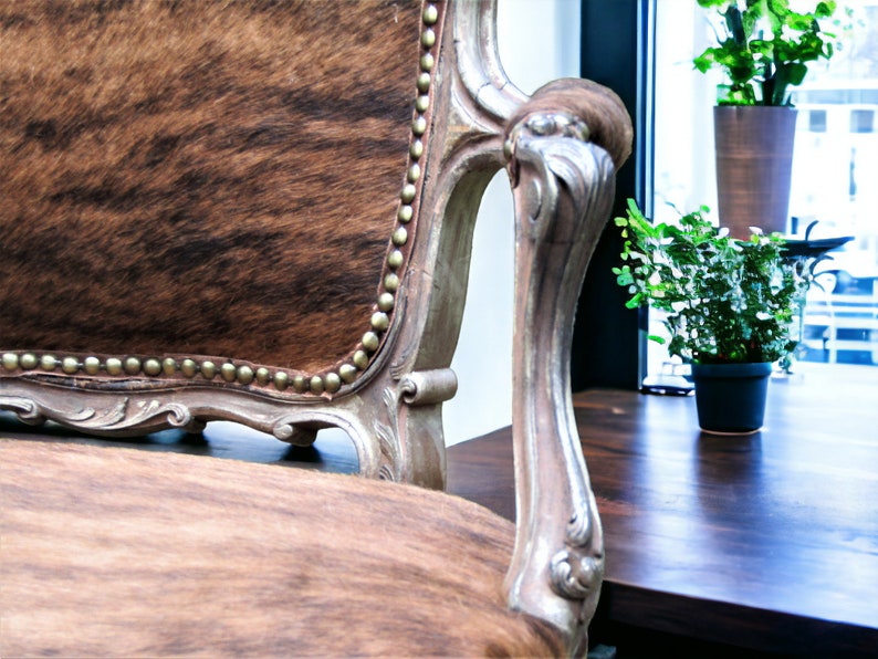 Pu&ograve; includere: Una poltrona in stile vintage con rivestimento in pelle di mucca marrone e bianca. La sedia ha una struttura in legno intagliato con finitura argentata e un bracciolo decorativo.