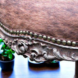 Pu&ograve; includere: Uno sgabello rivestito in pelle di mucca marrone con una cornice in legno decorata, ornata e dipinta in argento. La cornice ha una fila di chiodini in ottone attorno al bordo del sedile.