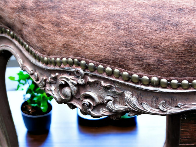 Pu&ograve; includere: Uno sgabello rivestito in pelle di mucca marrone con una cornice in legno decorata, ornata e dipinta in argento. La cornice ha una fila di chiodini in ottone attorno al bordo del sedile.