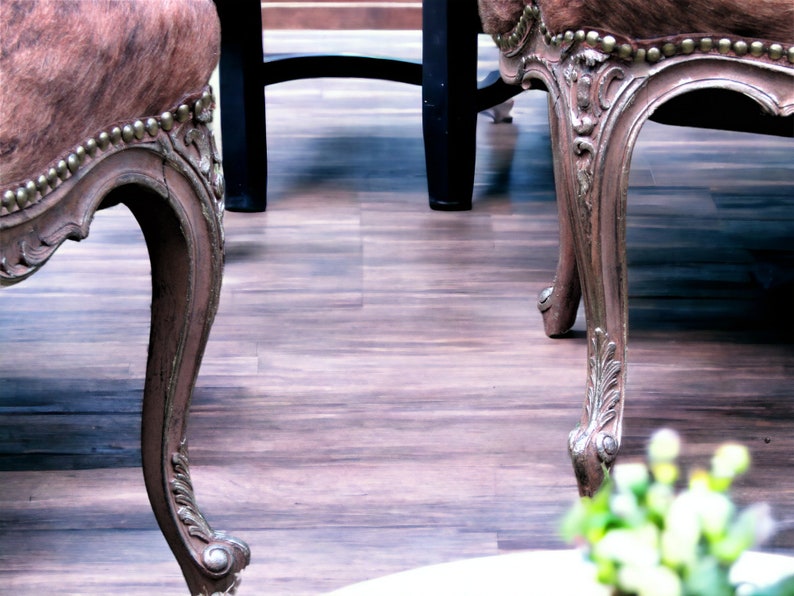 Pu&ograve; includere: Due sedie in legno ornate con rivestimento in pelle marrone e dettagli argentati. Le sedie sono posizionate su un pavimento in legno.