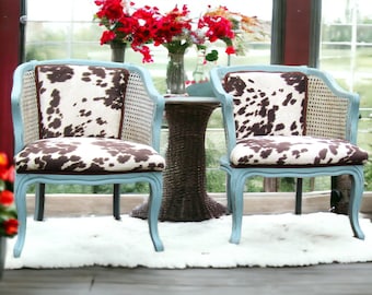 May include: Two blue chairs with a cowhide pattern upholstery, sitting on a white rug in front of a window. The chairs are facing each other and have a small round table between them.