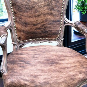 Pu&ograve; includere: Una poltrona in stile vintage con rivestimento in pelle di mucca marrone e bianca. La sedia ha una struttura in legno intagliato e dettagli decorativi.