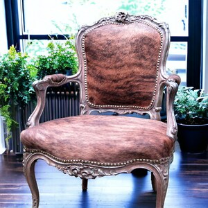 Pu&ograve; includere: Una poltrona in stile vintage con rivestimento in pelle di mucca marrone e bianca. La sedia ha una struttura in legno intagliato con dettagli decorativi e una finitura argentata.
