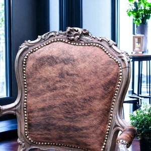 Pu&ograve; includere: Una poltrona in stile vintage con rivestimento in pelle di mucca marrone e bianca. La sedia ha una struttura in legno intagliato con dettagli dorati e uno schienale alto.