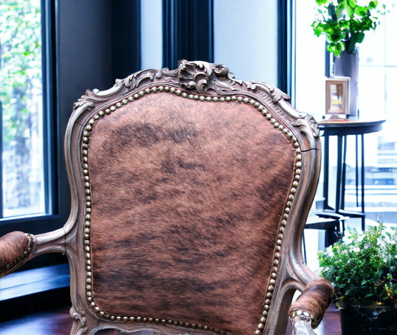 Pu&ograve; includere: Una poltrona in stile vintage con rivestimento in pelle di mucca marrone e bianca. La sedia ha una struttura in legno intagliato con dettagli dorati e uno schienale alto.