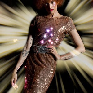 Pode incluir: Um vestido de lantejoulas bronze até o joelho com mangas curtas, decote redondo e uma faixa larga de lantejoulas cinza-escuras na cintura. A peça é coberta por lantejoulas metálicas refletoras que captam a luz, criando um efeito cintilante.