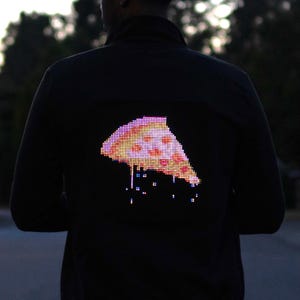 Może przedstawiać: Czarna kurtka z pikselowanym wzorem kawałka pizzy na plecach. Kawałek pizzy jest podświetlony różnymi kolorami, w tym czerwonym, żółtym i białym. Kurtka jest noszona przez osobę.