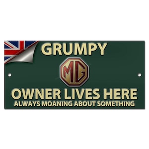 Puede incluir: Un letrero metálico verde oscuro con la inscripción dorada « GRUMPY OWNER LIVES HERE » y « ALWAYS MOANING ABOUT SOMETHING ». El letrero presenta el logotipo de MG y una bandera de la Union Jack en la esquina superior izquierda.