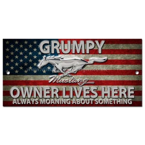 以下が含まれることがあります： アメリカ国旗のデザインが施された長方形の金属製サイン。「GRUMPY」、「MUSTANG」、「OWNER LIVES HERE」の文字が銀色で表示されています。中央には銀色のマスタングの馬のエンブレムがあります。