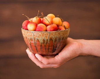 May include: A hand holds a brown and green ceramic bowl filled with ripe yellow and red cherries.