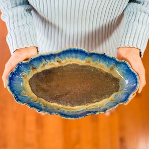 May include: A blue and brown ceramic serving dish with a wavy edge. The dish is oval shaped and has a dark brown glaze with a blue and yellow rim.
