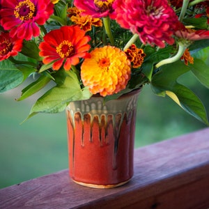 May include: A bouquet of bright red, orange, and yellow flowers in a red and green glazed ceramic vase. The vase has a rustic, handmade look.
