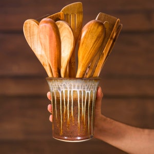 May include: A collection of wooden kitchen utensils, including spoons and spatulas, displayed in a ceramic utensil holder. The holder has a brown and cream glaze with a dripped design. The utensils are made of light-colored wood.