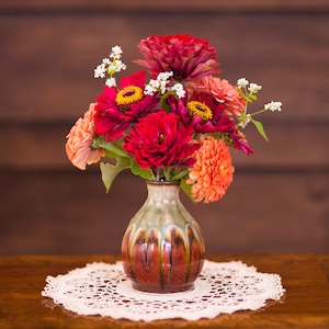 May include: A bouquet of red, orange, and white flowers in a brown and green ceramic vase. The vase has a textured surface and is sitting on a white crocheted doily.