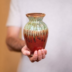 May include: A hand holds a small, red, green, and brown ceramic vase with a drip glaze.