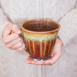 May include: A brown and green ceramic mug with a unique glaze pattern. The mug has a wide, rounded base and a slightly flared lip.
