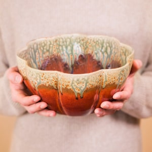 May include: A handmade ceramic bowl with a scalloped rim. The bowl features a gradient of colors, transitioning from a deep red at the base to a creamy beige at the rim, with green and brown accents. The bowl is held by two hands.