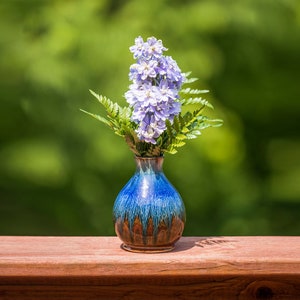 May include: A blue and brown ceramic vase with a textured glaze holds a bouquet of light purple flowers. The vase is sitting on a wooden surface.