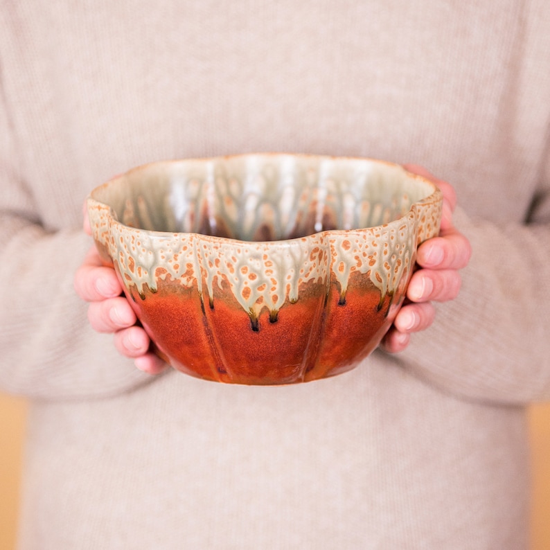 May include: A handmade ceramic bowl with a unique, scalloped rim. The bowl features a rich, reddish-brown base that transitions to a lighter, speckled tan and green at the rim. The glaze has a dripped effect, adding to its artisanal charm.