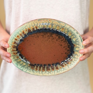 May include: A large oval ceramic serving dish with a green glaze and brown and black accents. The dish has a textured surface and is perfect for serving food.