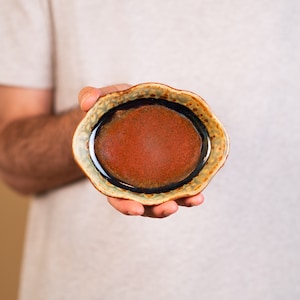 May include: A small, brown and black ceramic bowl with a wavy rim. The bowl has a red interior with a speckled glaze.
