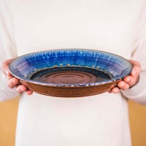 May include: A handmade ceramic plate with a wide rim. The plate features a rich, dark brown base, transitioning to a vibrant blue and purple glaze on the rim. The rim has a textured appearance with small, gold-colored accents. The plate is held by a person.
