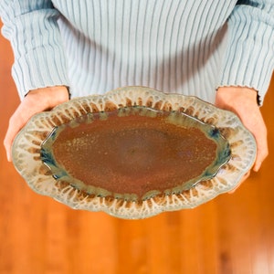 May include: A large, oval ceramic serving dish with a brown glaze and a white rim. The dish has a scalloped edge and a textured surface.