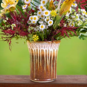 May include: A bouquet of white daisies, yellow irises, and red foliage arranged in a brown and white ceramic vase with a drip glaze.