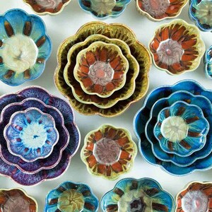 May include: A collection of ceramic flower-shaped bowls in various sizes and colors. The bowls feature a scalloped edge and a central design with shades of blue, brown, and cream. The arrangement showcases the bowls' unique shapes and artistic glaze.