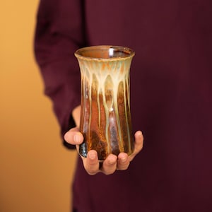 May include: A handmade ceramic vase with a unique glaze. The vase features a flared top and a cylindrical body. The glaze has a blend of colors, including brown, orange, and cream, with a drip effect. The vase is held in a person's hand.