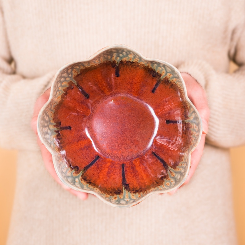 May include: A handmade ceramic bowl with a unique, scalloped edge. The bowl features a rich, reddish-brown interior with dark accents and a speckled, greenish-blue rim. The bowl is held by a person wearing a cream-colored sweater.