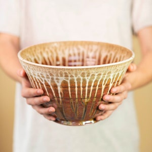 May include: A handmade ceramic bowl with a unique glaze. The bowl features a warm color palette of brown, beige, and white, with a dripped glaze effect. The bowl is held by a person, showcasing its size and shape. A decorative piece for the kitchen.