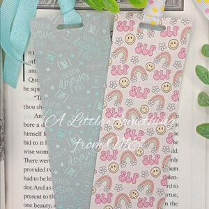 May include: Two decorative bookmarks with ribbon ties. One is light gray with the text "EVERYONE deserves a VOICE" in a repeating pattern. The other is pink with rainbows, smiley faces, flowers, and the letters "SLP".