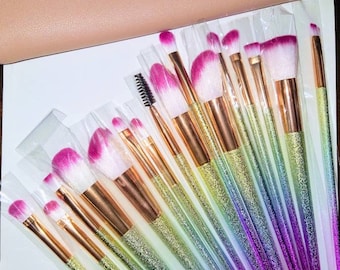 May include: A set of 20 makeup brushes with rainbow glitter handles. The brushes are individually packaged in clear plastic sleeves. The handles are a gradient of colours, starting with yellow and transitioning to purple.