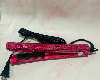 May include: A vibrant pink flat iron, encrusted with small, glittering rhinestones. The hair straightener features black plates and a black power cord with a three-pin plug. It rests on a light pink and white surface.