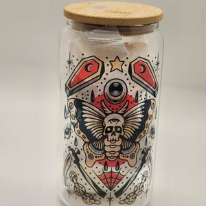May include: A glass jar with a bamboo lid featuring a colorful tattoo-style design. The design includes a skull with an eye, a butterfly, a spider web, and other symbols.