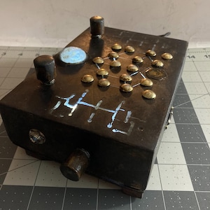 May include: A rectangular black electronic device featuring a grid of brass-coloured metal studs, two dark cylindrical knobs, a circular light-up element, and a hand symbol icon. The surface displays etched white markings and a side-mounted input jack.