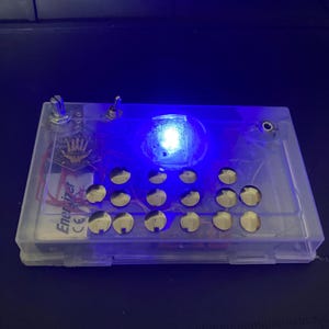 May include: A rectangular, translucent plastic electronic device enclosure containing internal circuitry, an Energizer battery, and a grid of fifteen circular metal buttons. A bright blue LED light is illuminated on the top surface, which also features two metal toggle switches.