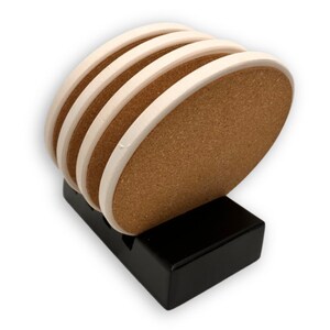 May include: Set of five oval coasters with a natural cork center and a cream-colored border. The coasters are stacked in a black, rectangular holder. The coasters are designed to protect surfaces from drink rings.