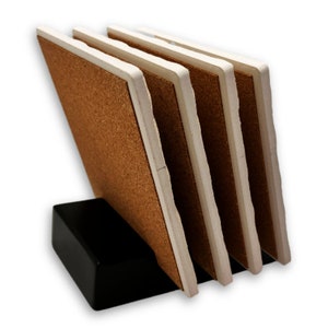 May include: A set of four square coasters with a cork surface and white edges, displayed in a black holder. The coasters are angled, showcasing their brown and white color scheme. The holder is a solid black rectangle.